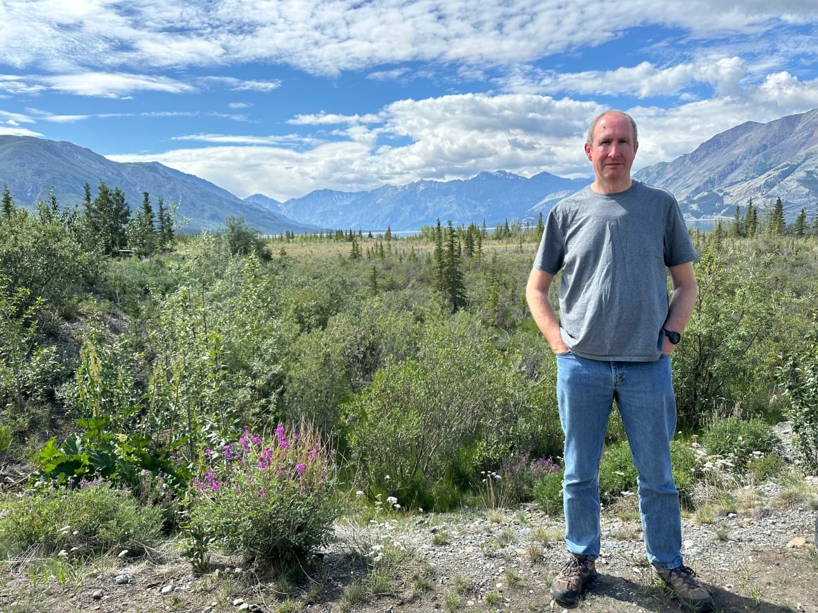A man stands outside, with a wide mountain valley stretching behind him.