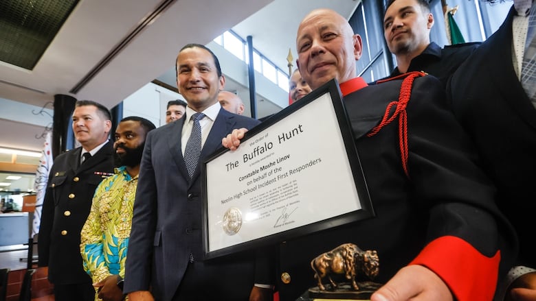 A man in uniform smiles while holding a statuette of a buffalo and a framed document that says in big letters 'The Order of the Buffalo Hunt' and 'Constable Moshe Linvo on behalf of the Neelin High School Incident First Responders