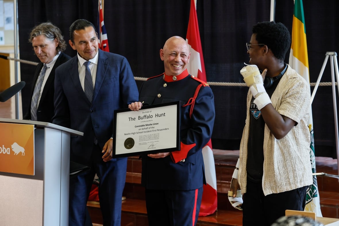 A man in uniform and a man in a soot stand in front of a podium. Both are looking toward a teenager who has his both hands bandaged. The man in the uniform is holding a framed document, and is smiling.