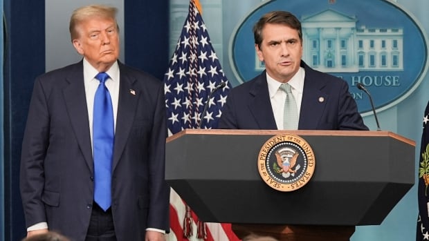 A cleanshaven man with dark hair in a suit and tie speaks at a podium in front of an American flag. Another, older cleanshaven man in suit and tie looks on.