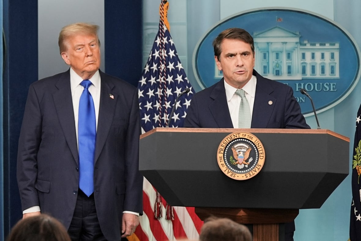 A cleanshaven man with dark hair in a suit and tie speaks at a podium in front of an American flag. Another, older cleanshaven man in suit and tie looks on.