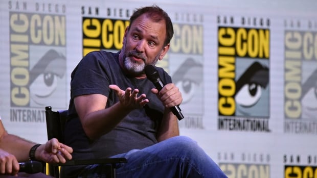 A man in jeans and a black shirt shrugs while holding a microphone