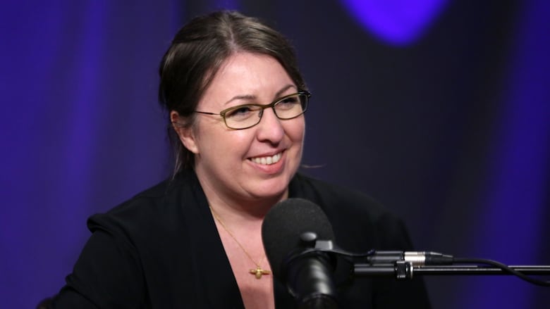 A smiling woman wearing glasses sits in front of a studio microphone.