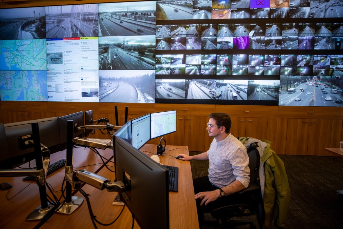 A man sits at a computer inside a large room with multple screens on the wall.