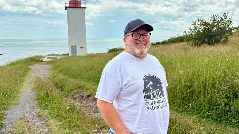 A man in a tee shirt stands in front of a lighthouse and the ocean