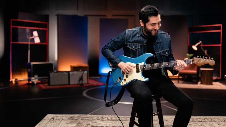 A man with short dark hair sits on a stool playing a blue electric guitar in a studio.