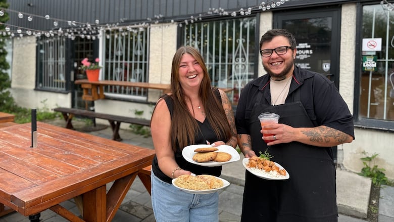 A woman with long hair and a man with dark black glasses stand outside with plates of food.