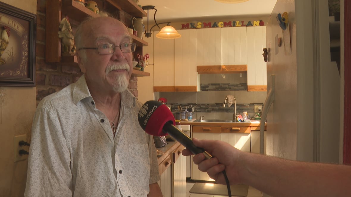 A man wearing a white shirt sits and talks into a CBC News microphone.
