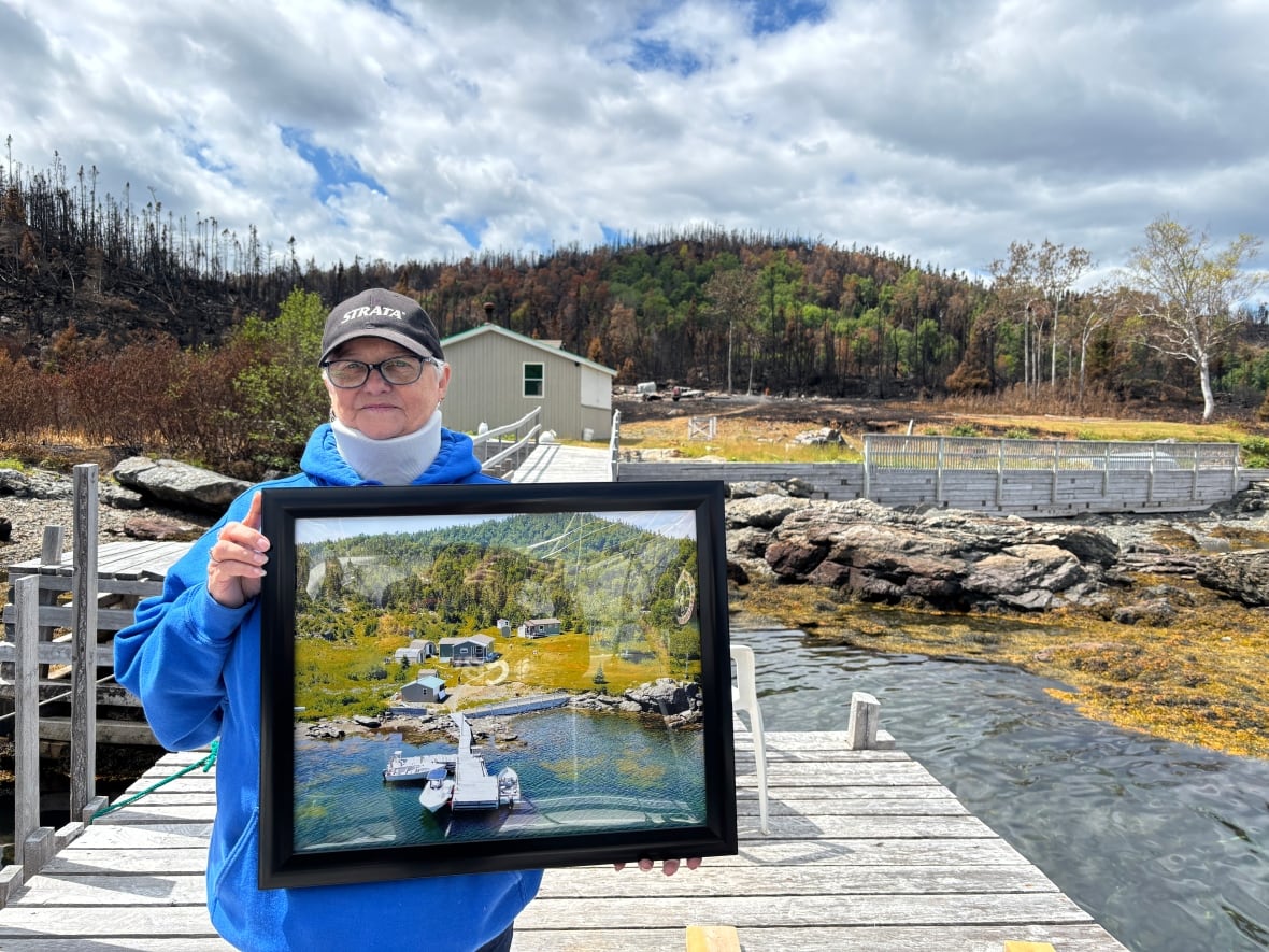 A woman wearing a blue hoodie and baseball cap holds a large photograph, while standing on a dock in front of a cabin that has been destroyed by a wildfire.