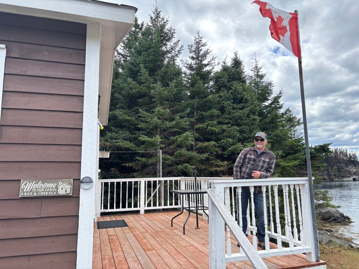 A man wearing a plaid jacket and sunglasses leans against the railing of the deck attached to his cabin.