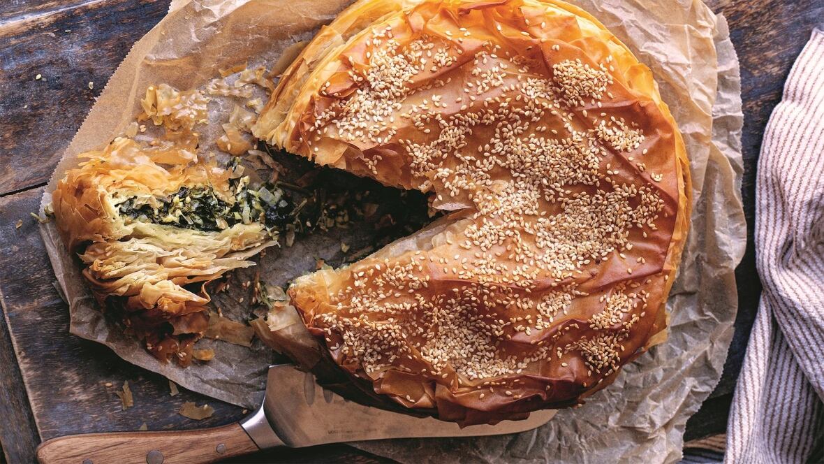 Top down shot of a sesame seed-topped phyllo spinach pie on a wooden surface. A slice is cut from it and sitting next to a knife. 