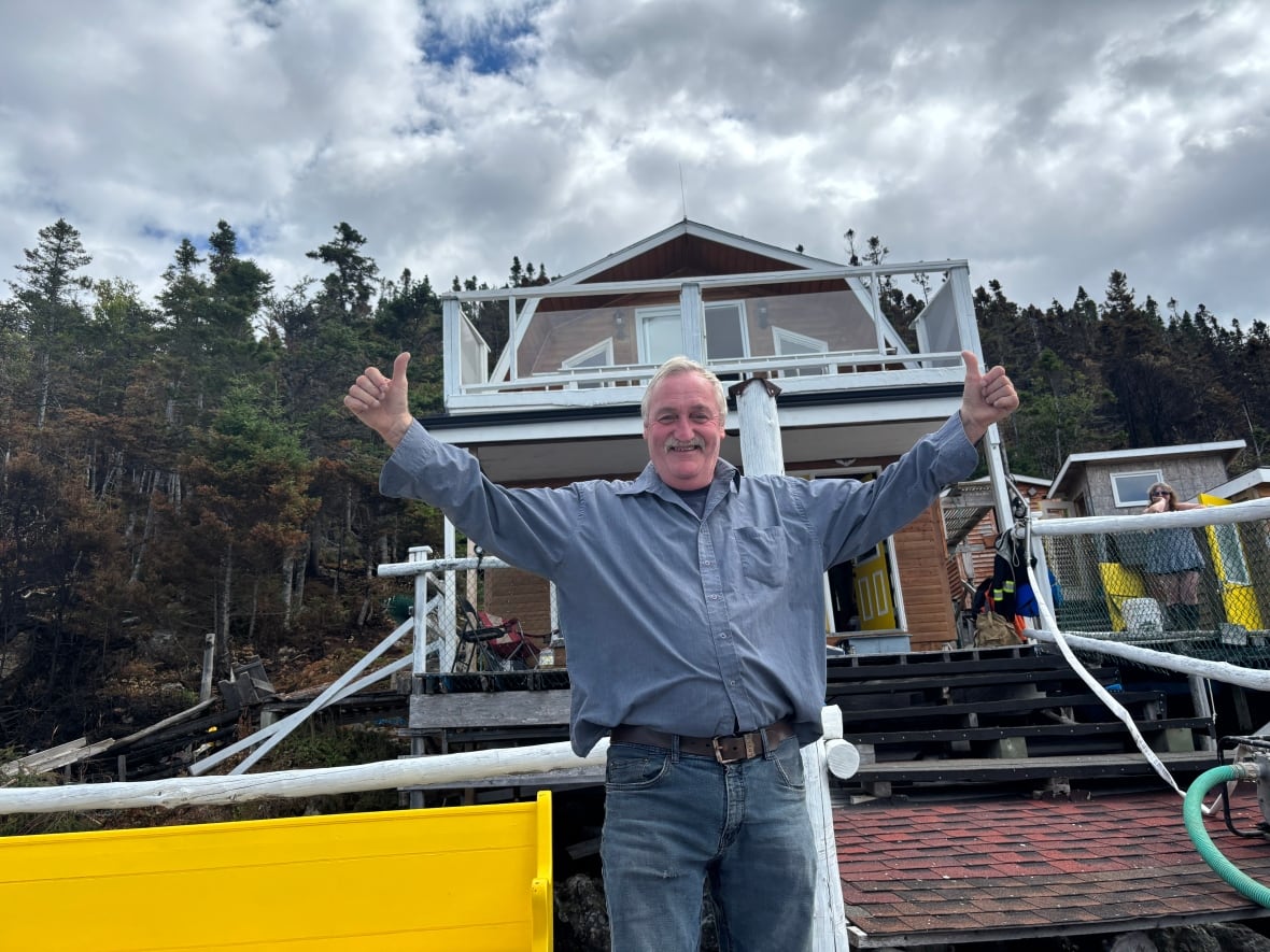 A man wearing jeans and a button up shirt puts both his thumbs up while standing in front of a cabin.