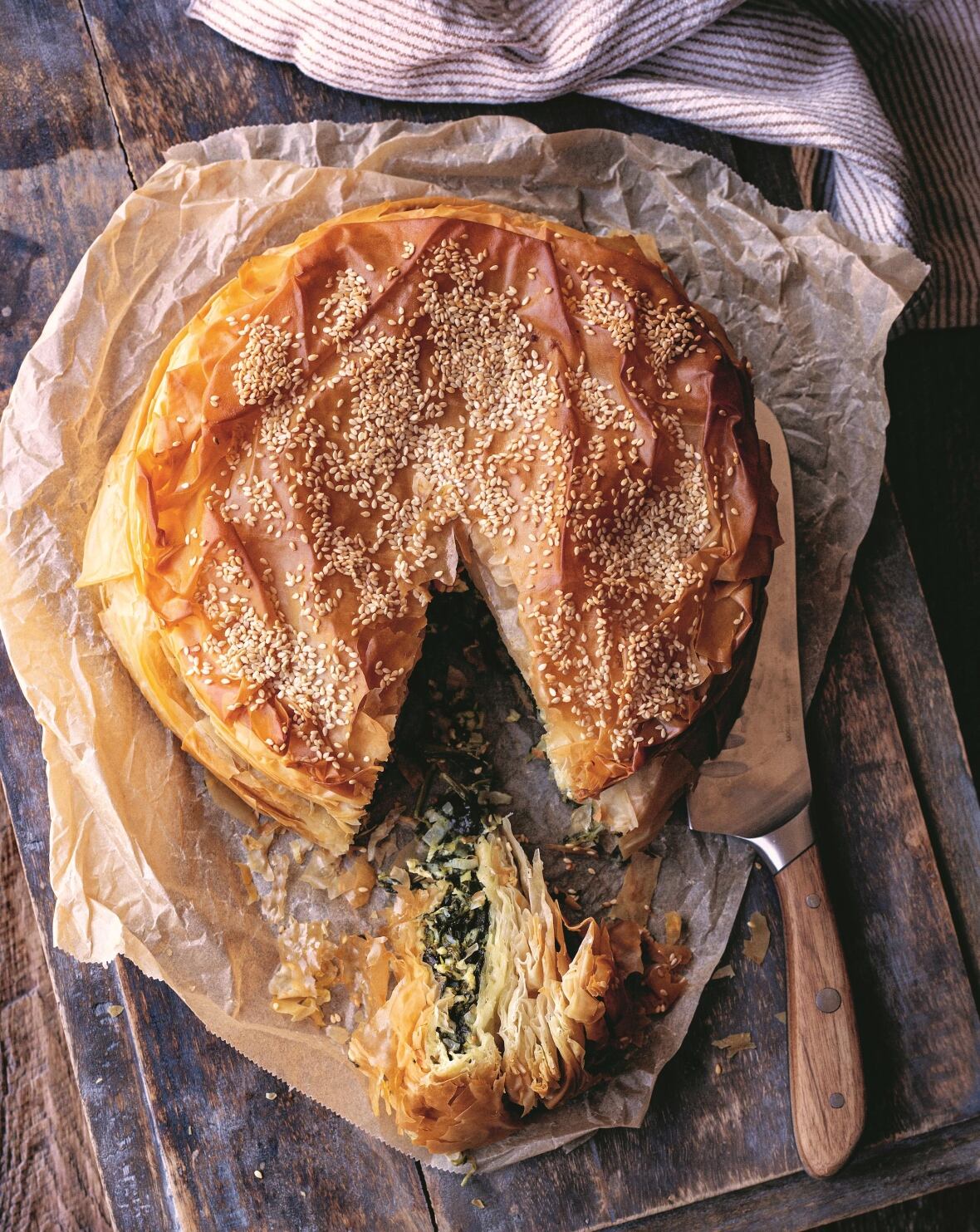 Top down shot of a sesame seed-topped phyllo spinach pie on a wooden surface. A slice is cut from it and sitting next to a knife. 