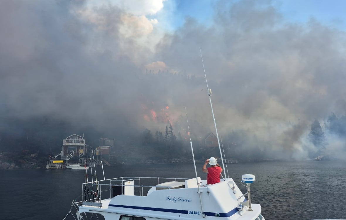 A man on a boat looks at flames and smoke on a forested shore, as a cabin sits nearby to the left of the flames.