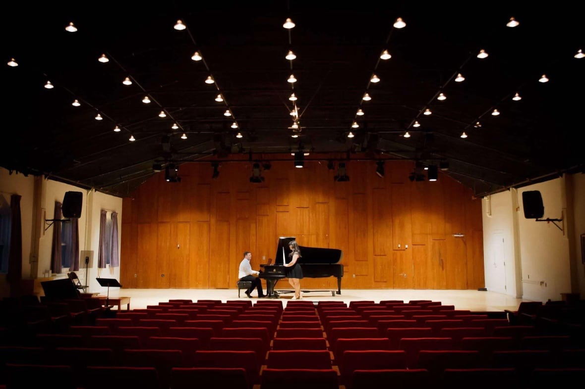 Two people standing at a piano on the stage of an empty recital hall.