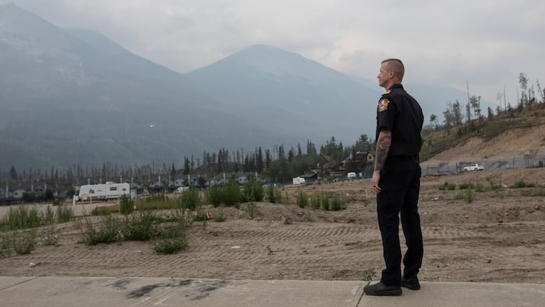 A man in a black uniform overlooks a barren landscape that has been devastated by wildfire. Mountains with clouds around them are seen in the distance.