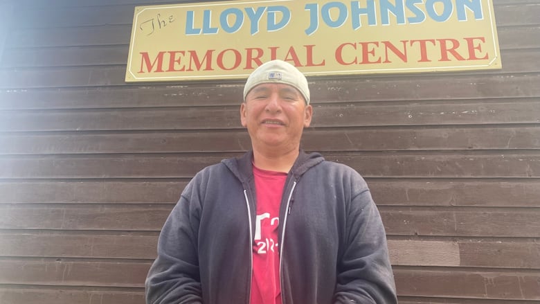 A man in a hoodie and a red shirt and a ball camp stands under a sign reading Lloyd Johnson Memorial Centre.