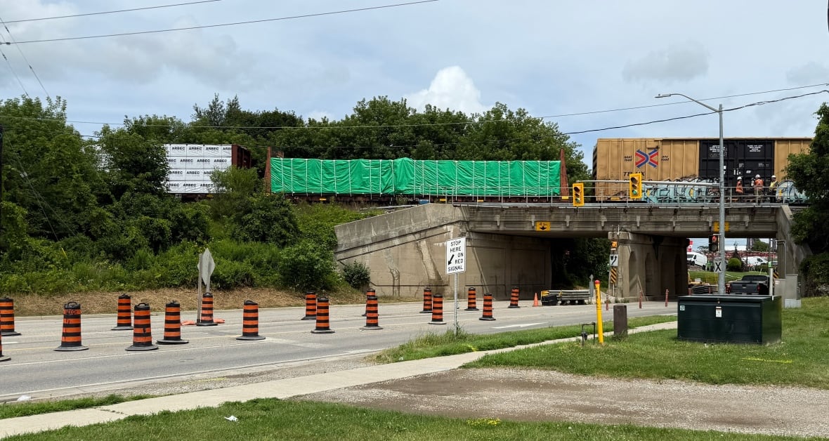Uma rua com torres de construção e uma ponte sobre a rua com um trem parado na ponte e trabalhadores com coletes de segurança estão ao lado do trem