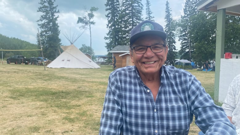 A man in glasses and a blue-checked shirt can be seen with a teepee behind him.