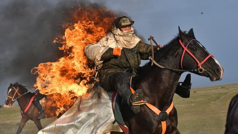 A man rides a horse with a blanket on fire on its rear in a sunny field.