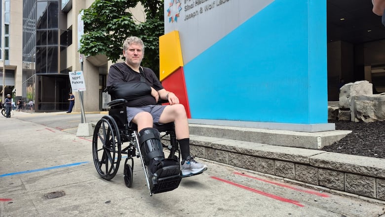 A man with a cast on his leg and his arm on the right side sits in a wheelchair on a sidewalk.