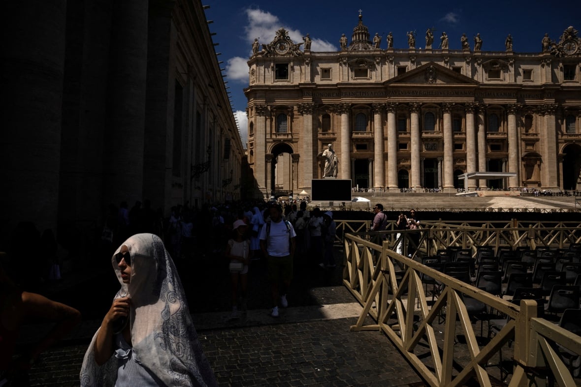 A woman holds a white scarf over her head while a large stone building is illuminated by sunlight.