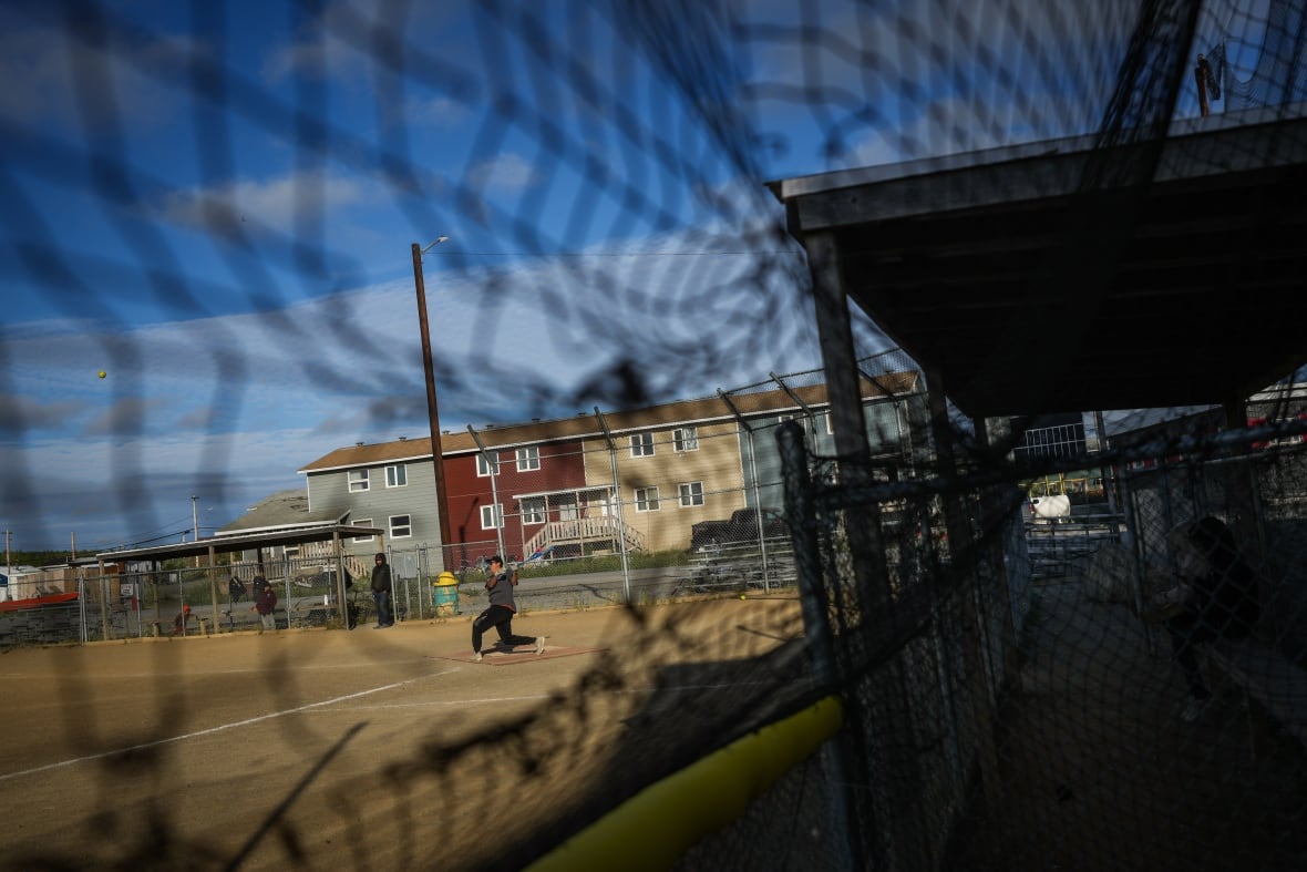 A man swings a baseball bat on a dusty field seen through a hole in a metal fence.