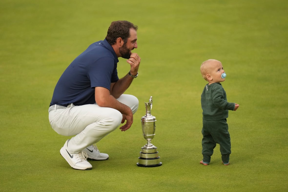 A man kneels on green grass near a silver trophy as a young toddler in green stands looking away.