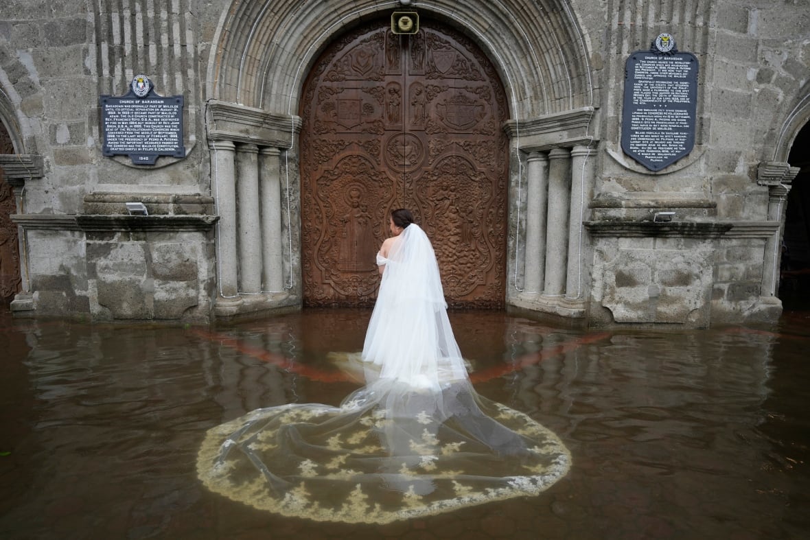 A woman in a white wedding dress stands in front of a large stone building while in murky brown water.