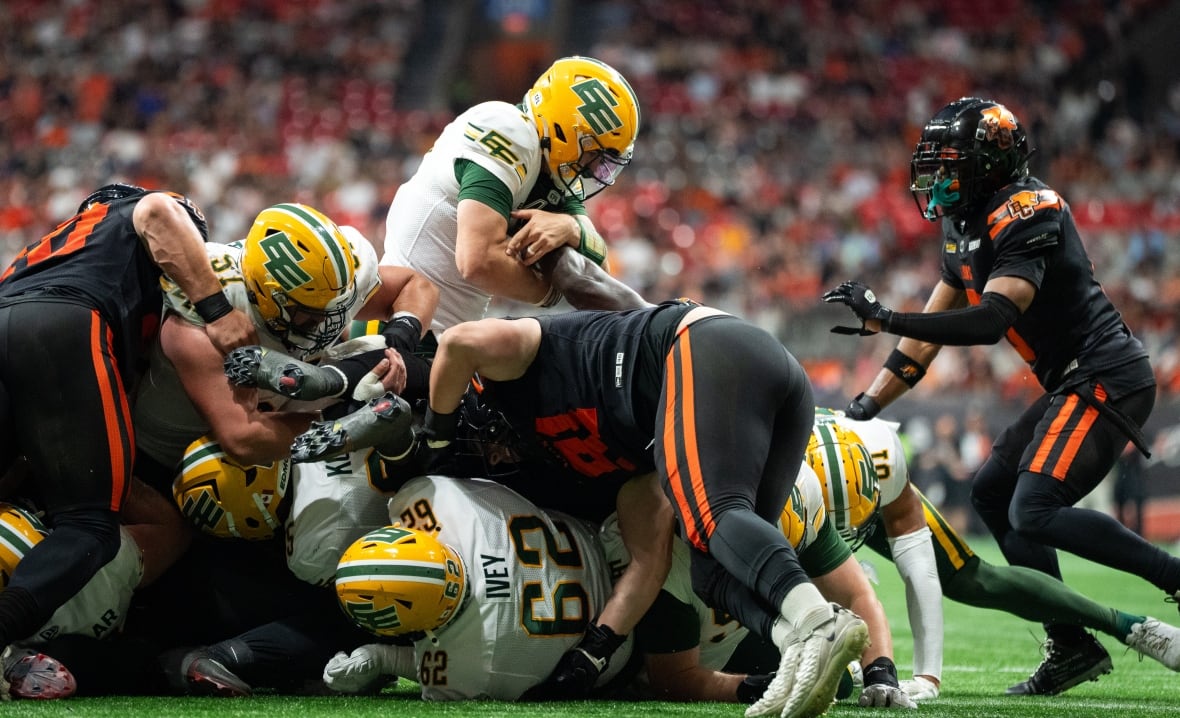 A quarterback in a white, yellow, and green jersey attempts to jump over a scrum of football players. Some of the players are in white, yellow, and green jerseys, and the other half are in orange and black jerseys.