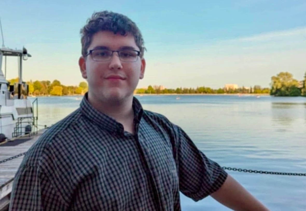A young man with curly black hair and a collared shirt smiles at the camera while standing on a dock with water in the background.