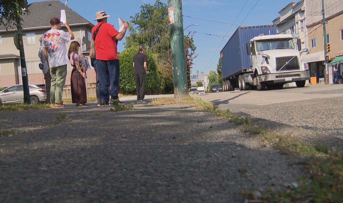 Standing on a sidewalk, a group of six people raise signs in protest as a container truck drives by them.