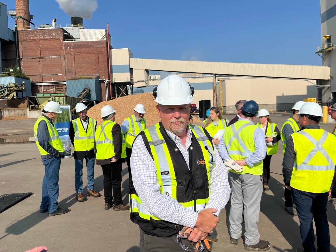 A man in a high-vis vest stands in front of a crowd of people in green high-vis vests at a industrial manufacturing plant