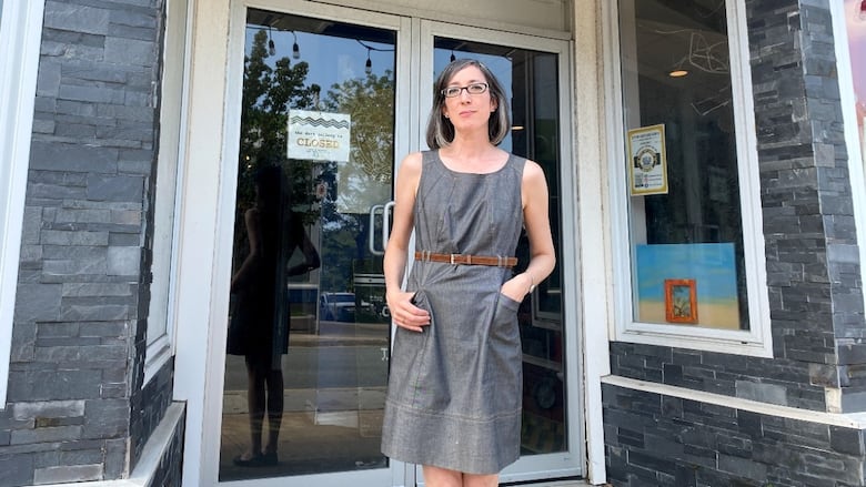 A white woman with a dark-grey bob and glasses wears a grey knee-length dress, standing outside glass doors of a building