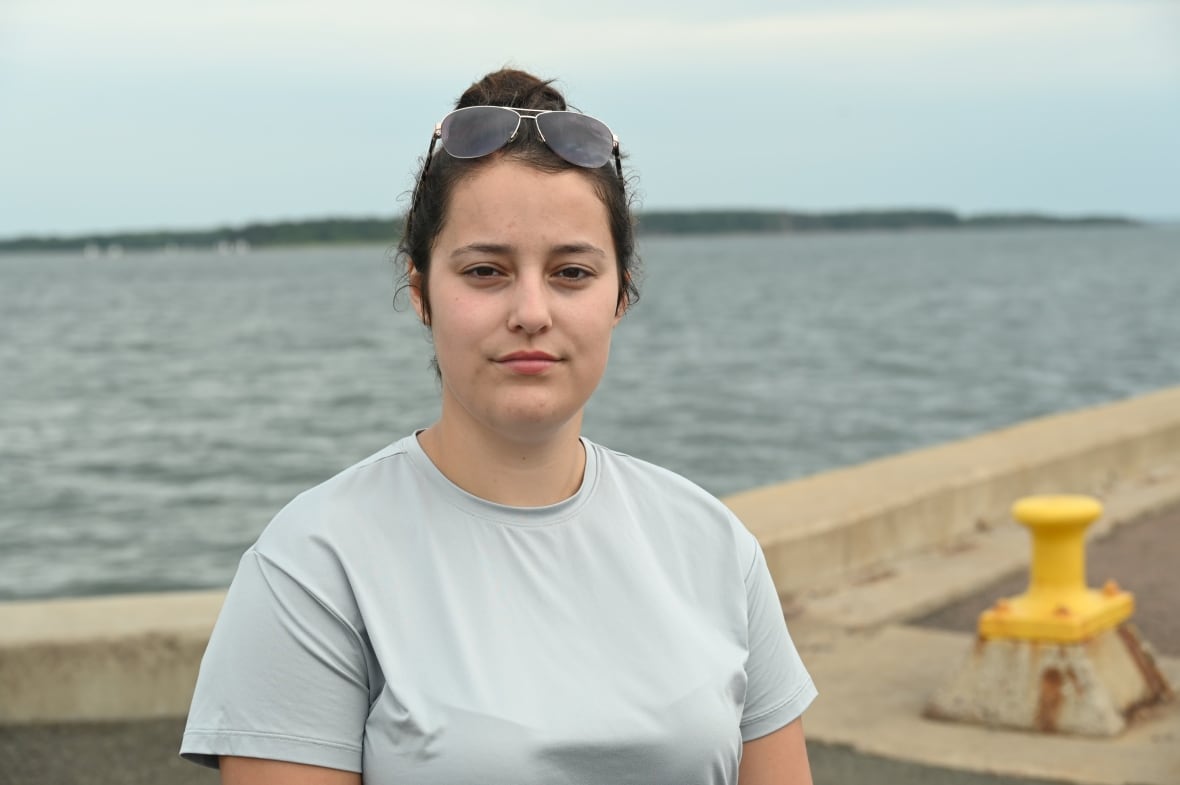 A young woman with a grey shirt and her hair in a bun, standing in front of water.