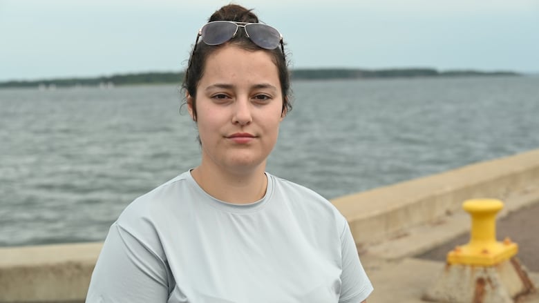 A young woman with a grey shirt and her hair in a bun, standing in front of water.
