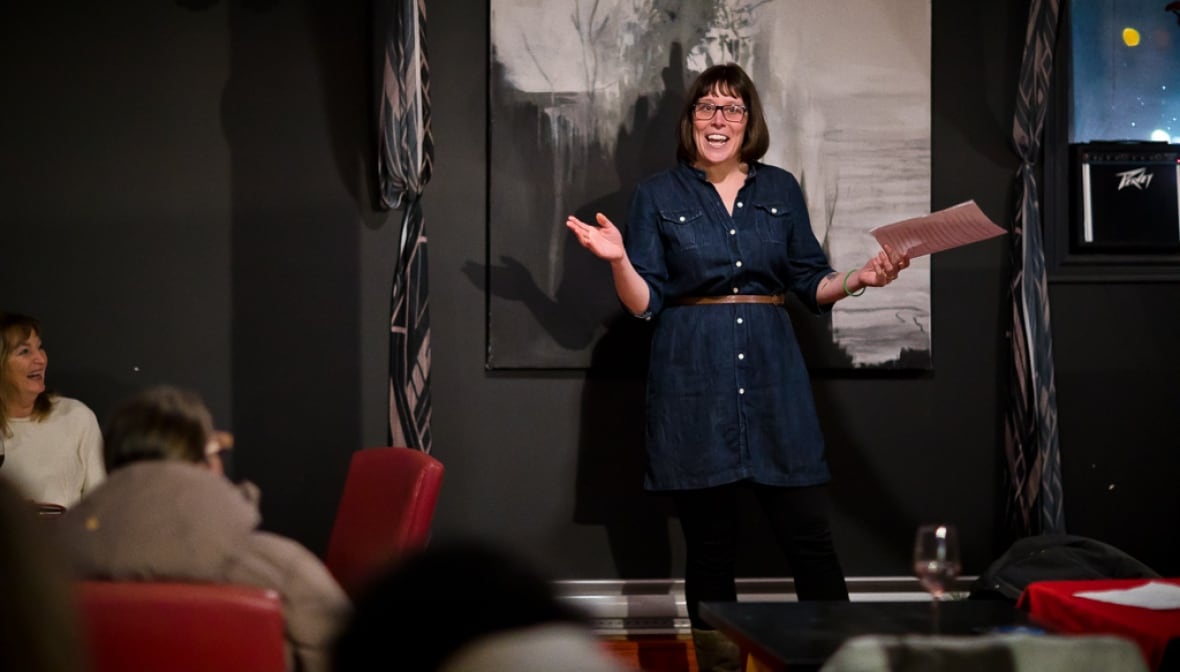 A white woman with glasses and brown hair in a bob wears a navy denim dress and brown belt, holding papers in one hand. She stands on a stage in front of tables of people in red chairs