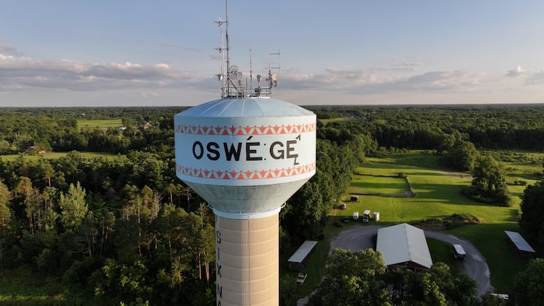 A tall water tower marked with the word “OSWE:GE̲” rises above green fields and forests in Ohsweken, Six Nations of the Grand River.
