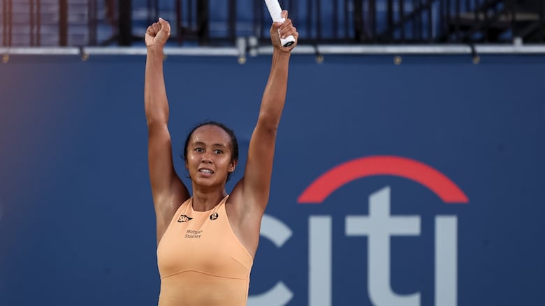 A female tennis player raises both arms straight up over her head, forming fists with her hands.
