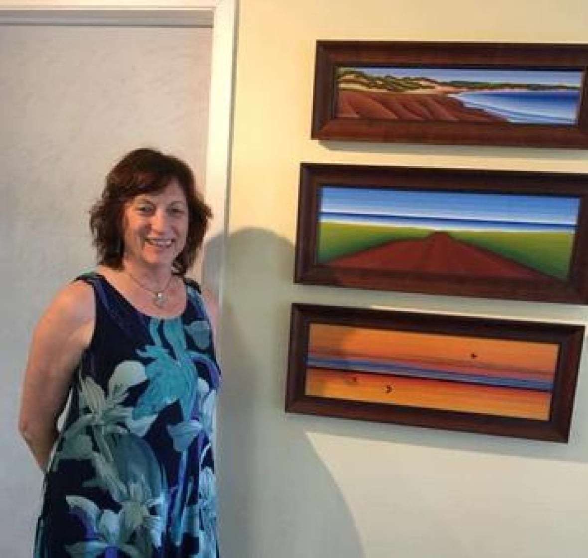A woman in a blue flower print dress stands in front of three paintings of P.E.I. landscapes.