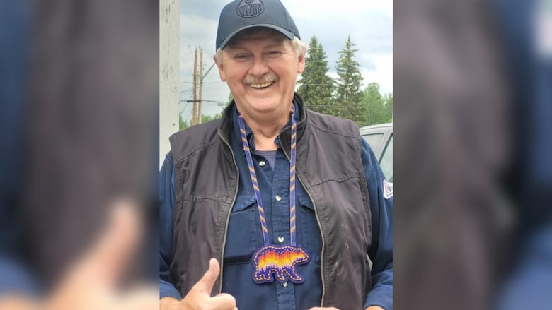 A man in a hat, wearing a dark sweater and vest poses with a beaded bear necklace he was gifted from a community member.