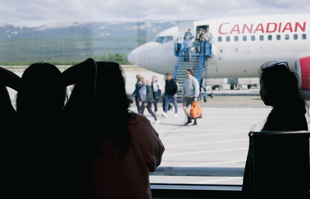 A few silhouettes stand in front of the airport window as a plane unloads with passengers.