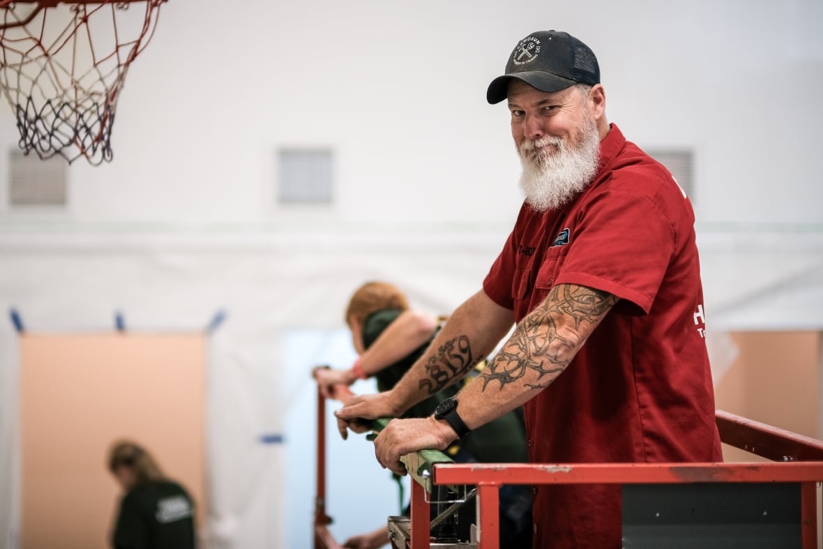 A man with white beard, black cap and red T-shirt looks at the camera while standing on a cherry-picker platform.