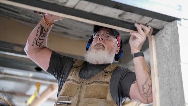 A man with white beard, wearing brown overalls, installing a window frame in a new building.