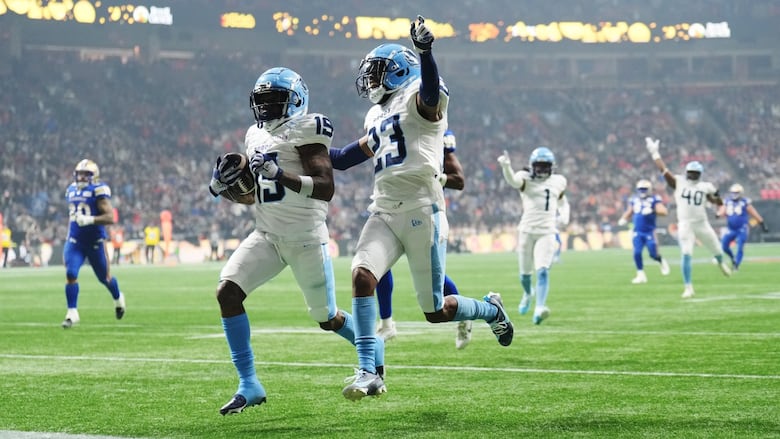 A player in blue and white carries the ball into the end zone, while a teammate runs alongside, his arm raised in celebration.