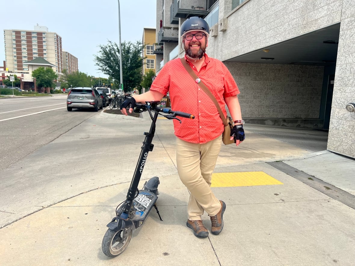 A man leans on his electric scooter, he is smiling towards the camera wearing a helmet, and his legs are crossed.
