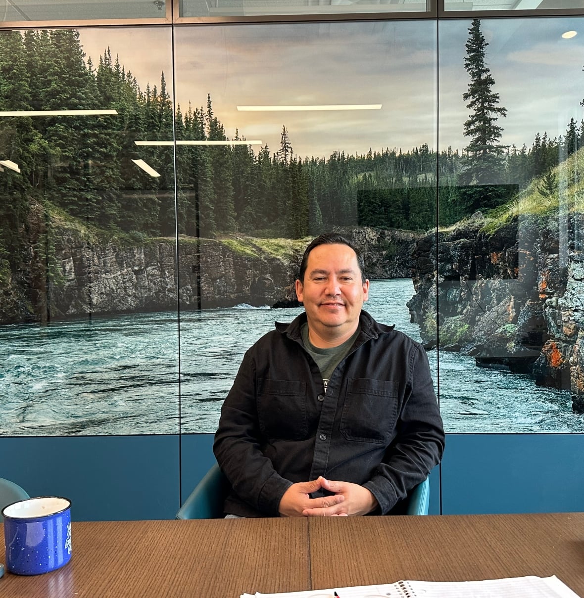 A man sitting at a table with his hands crossed and a photo of a river in the background.