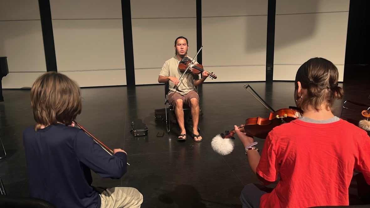 A man playing fiddle on a stage, with some children playing violin in the foreground.