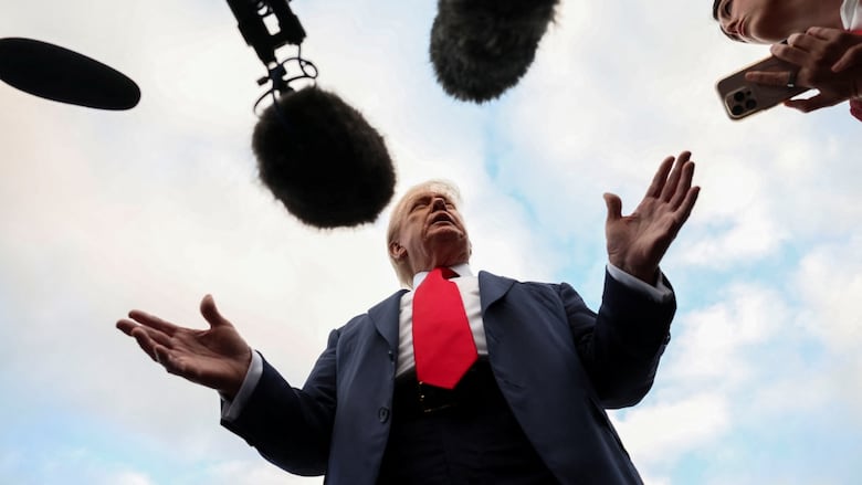 An older man wearing a navy suit and a red tie gestures as he stands in front of microphones.