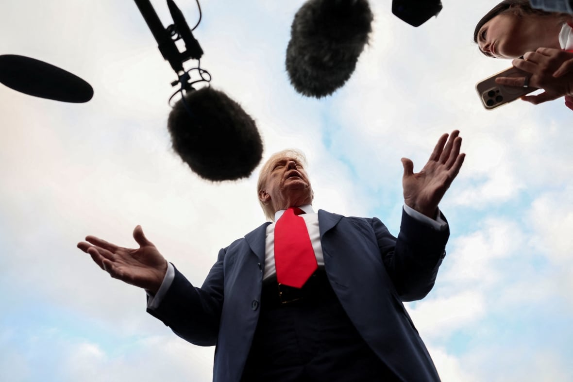 An older man wearing a navy suit and a red tie gestures as he stands in front of microphones.